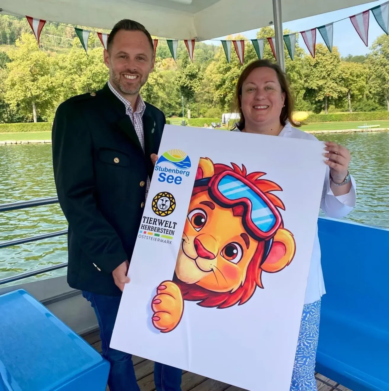 A man and a woman are standing on a boat, holding a poster with a lion wearing goggles. They are smiling and posing for a photo. The boat is on a lake with trees and mountains in the background.