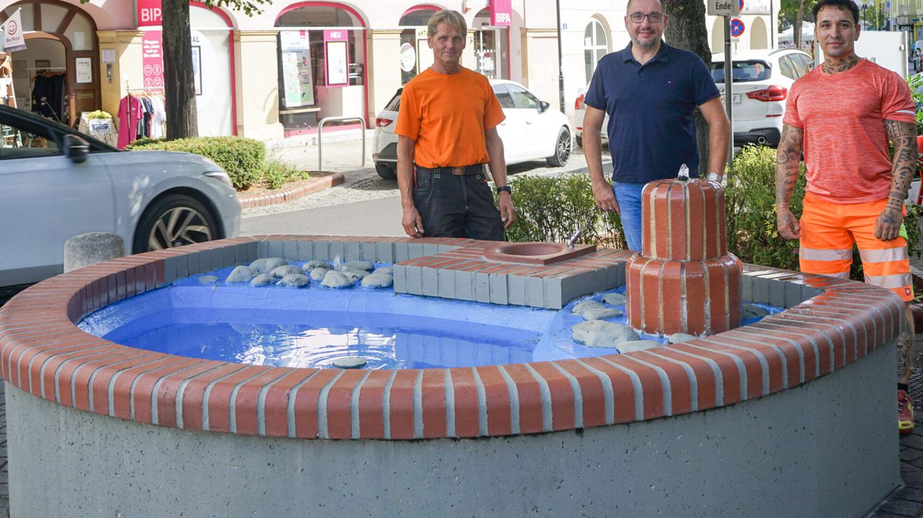 Two men stand next to a fountain in a city square, with a building and parked cars in the background.