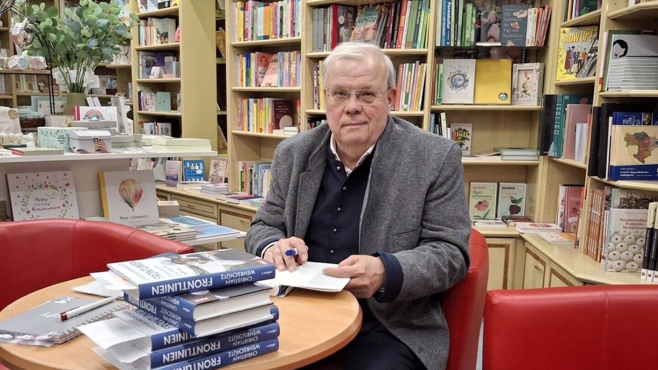 Ein älterer Mann mit Brille sitzt in einer Buchhandlung an einem Tisch, umgeben von Büchern, und scheint zu schreiben oder zu lesen.