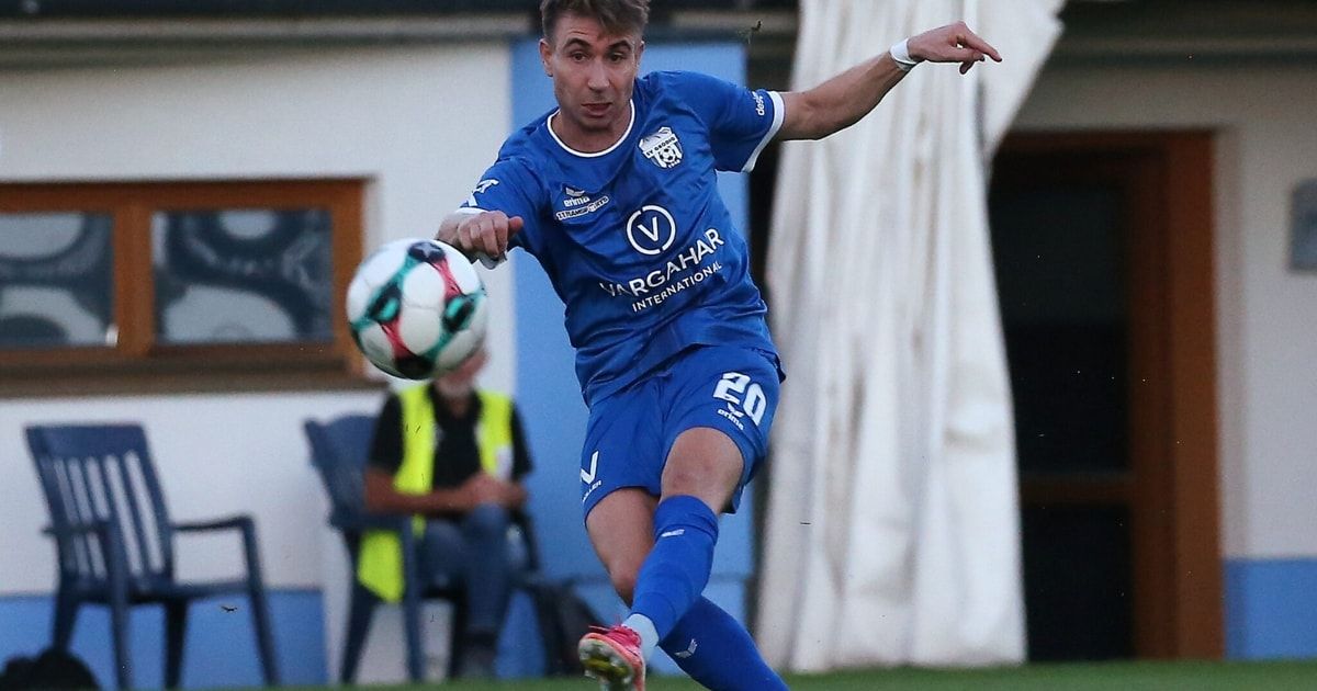 A soccer player in a blue jersey with the number 20 on it is kicking a soccer ball. In the background, a person is sitting on a chair wearing a yellow vest.