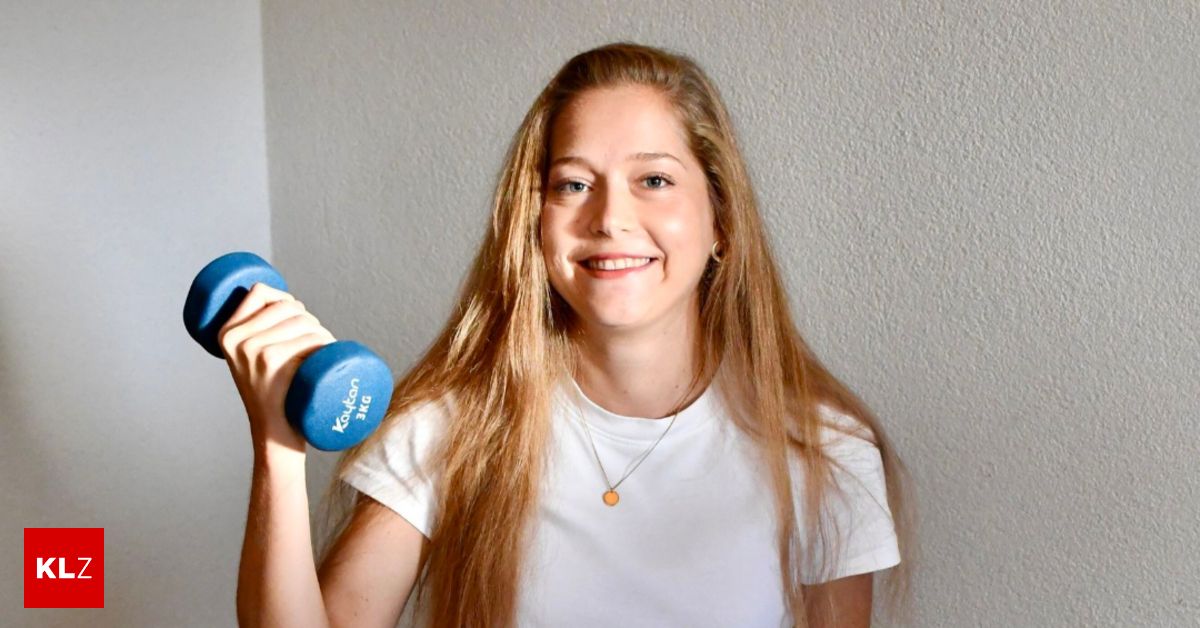 Eine Frau mit langen blonden Haaren lächelt und hält eine blaue Hantel in der rechten Hand. Sie trägt ein weißes T-Shirt und eine Halskette.