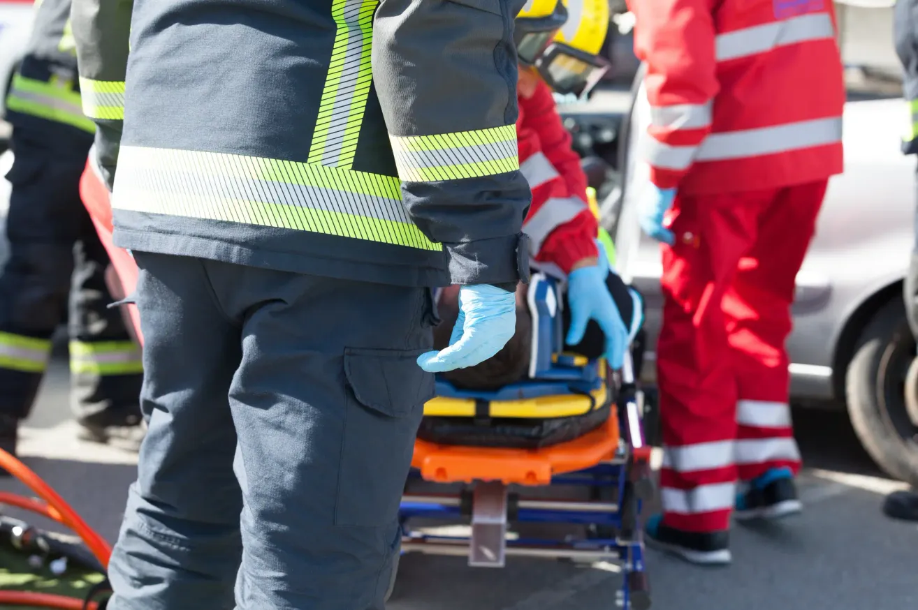Rettungskräfte in Schutzkleidung transportieren einen Patienten auf einer Trage. Ein Helfer trägt einen gelben Helm und Handschuhe.