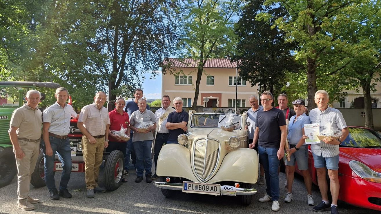 A group of men stand around a vintage car with baskets on its hood, posing for a photo in front of a building.