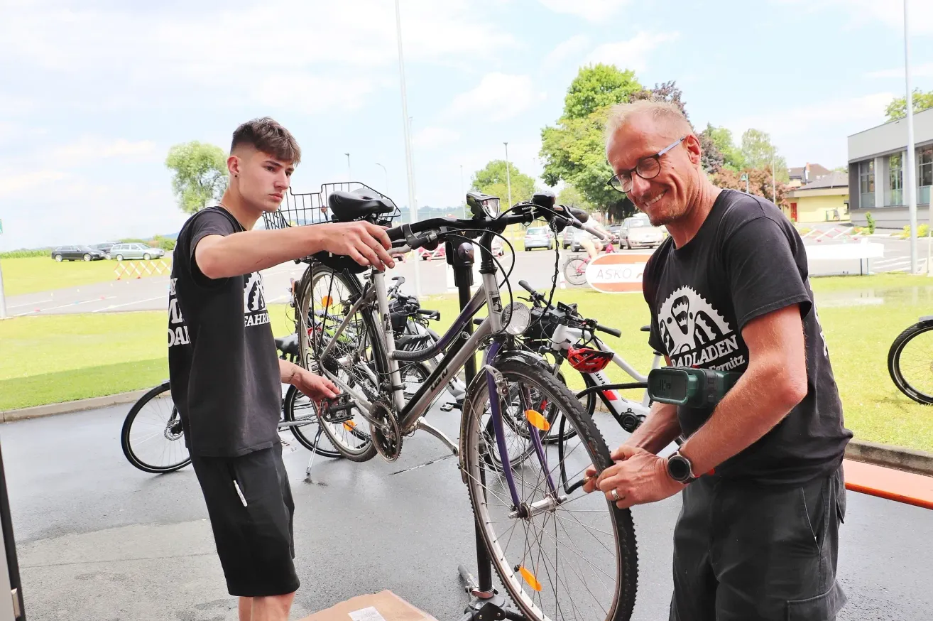 Two men are fixing a bicycle in an outdoor setting. One is holding the bike while the other is adjusting the front wheel. They are both wearing black t-shirts.