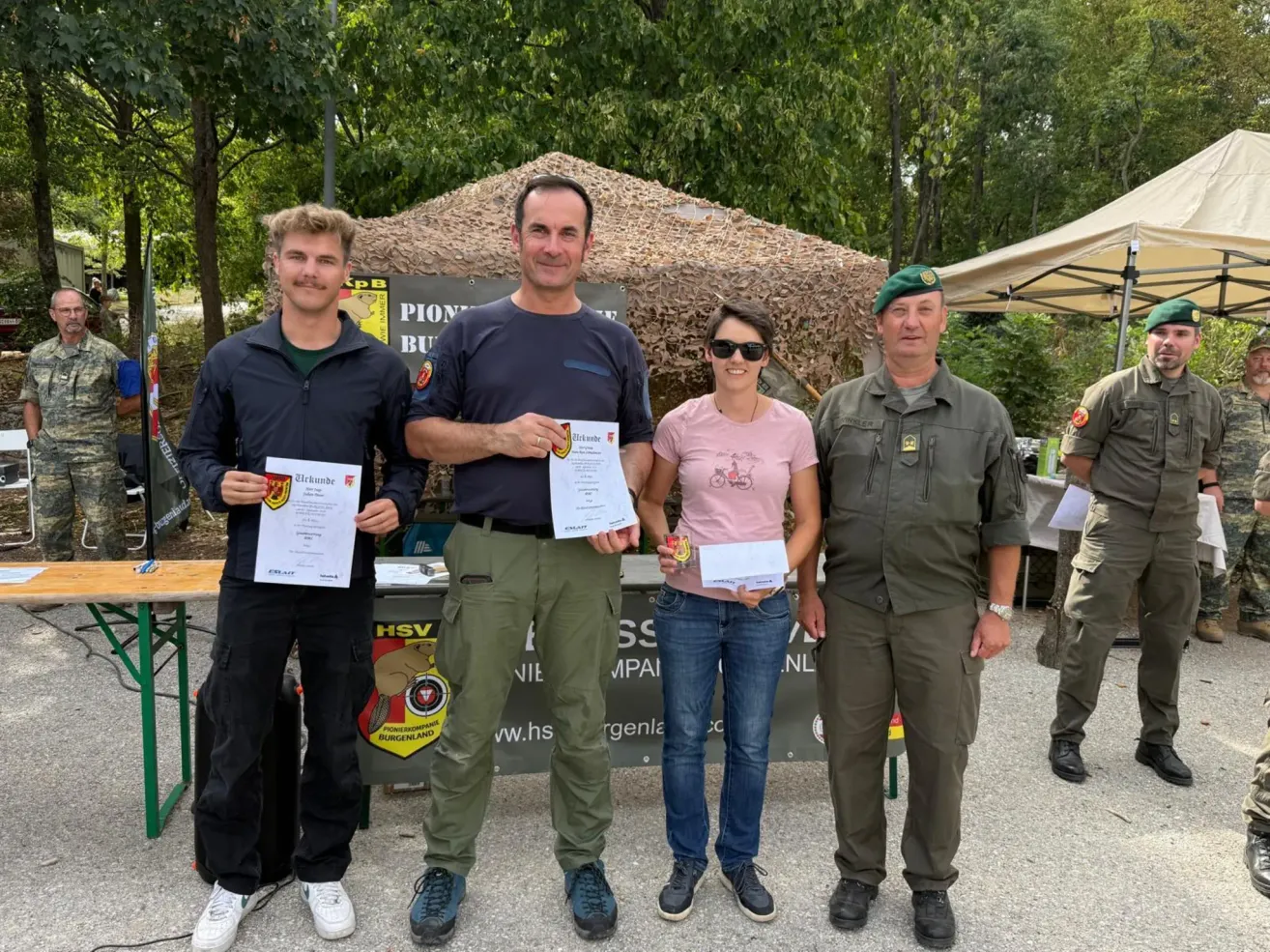 Vier Personen stehen zusammen und halten Urkunden. Sie sind in verschiedenen Uniformen gekleidet und scheinen sich bei einer Veranstaltung im Freien zu befinden. Hinter ihnen steht ein Zelt und ein Tisch mit einem Schild.