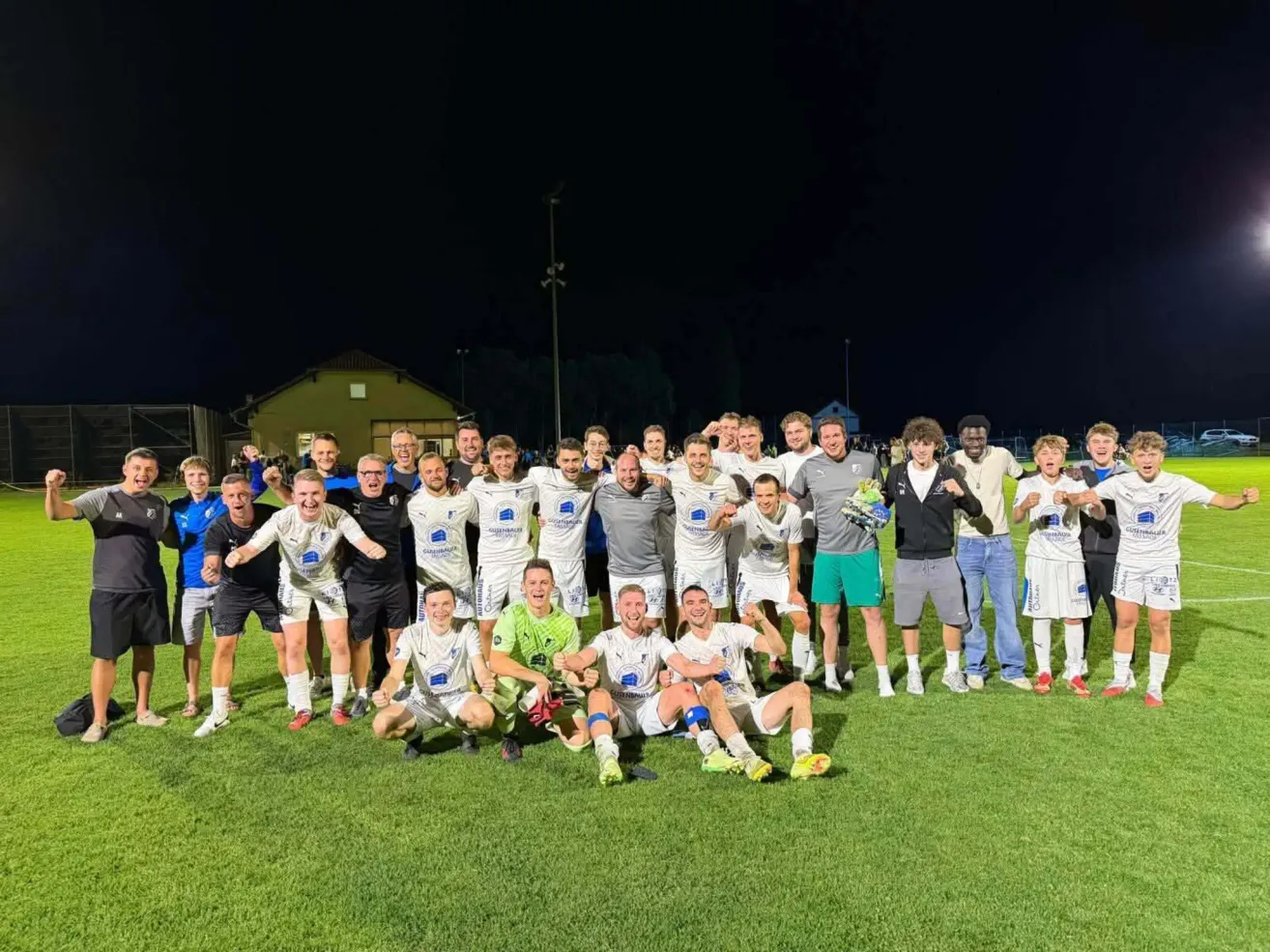 A soccer team poses for a group photo on a field at night, with players in white uniforms and some in casual clothes.