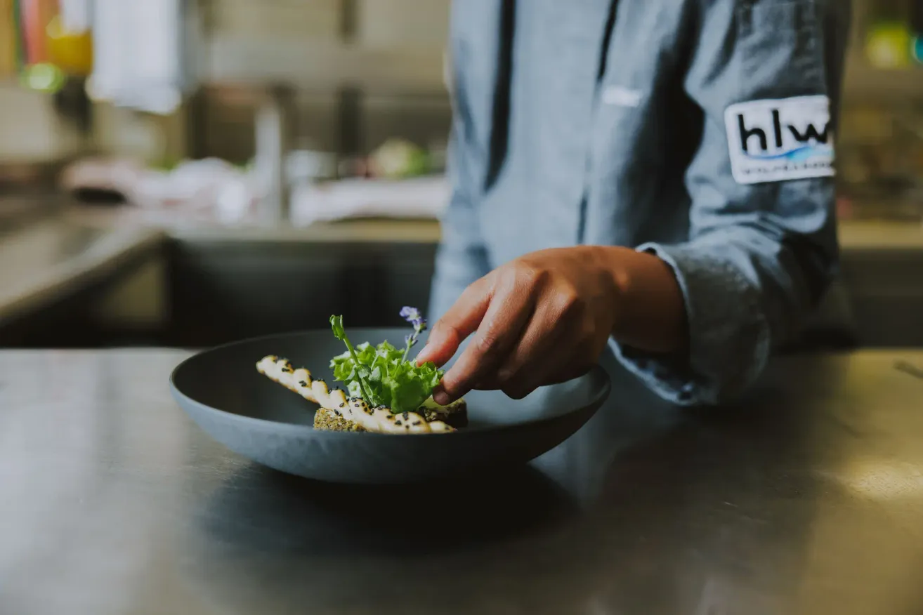 A chef in a kitchen, placing a garnish on a plate with a dish. The plate is on a countertop.