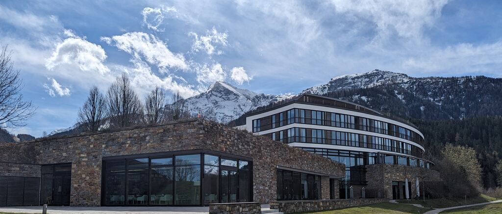 A modern building with glass walls and stone facade, set against a backdrop of snowy mountains and a blue sky with clouds.