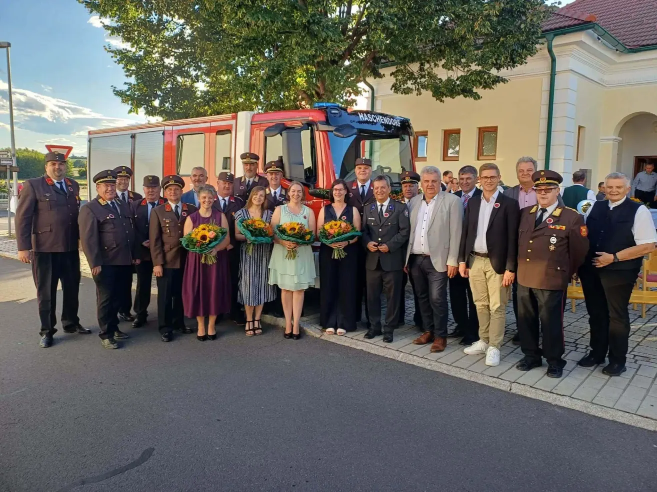 Eine Gruppe von Menschen in Uniform und formeller Kleidung steht vor einem roten Feuerwehrauto und hält Blumensträuße.
