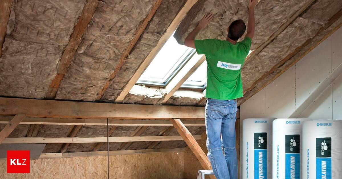 A man in a green shirt is installing insulation in an attic with a skylight. He is standing on a ladder.
