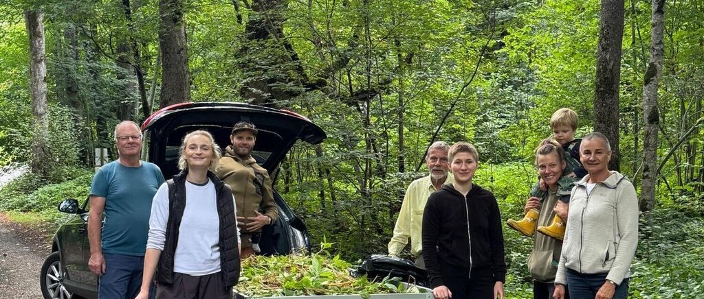A group of people, including adults and children, are standing in a forest. They are smiling and appear to be posing for a photo. A car with its trunk open is behind them.
