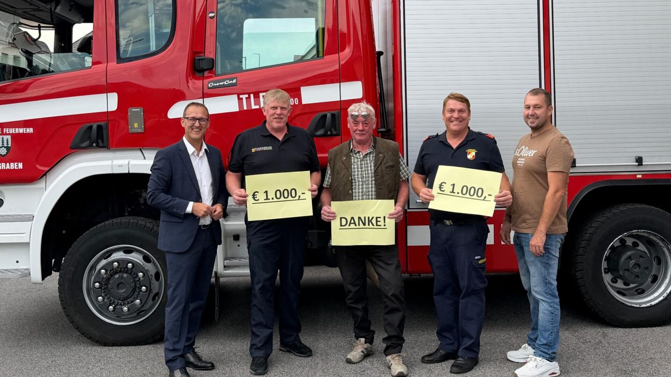 Fünf Männer stehen vor einem Feuerwehrauto und halten Schilder mit '€1.000,-' und 'DANKE!' Zwei der Männer tragen Uniformen.