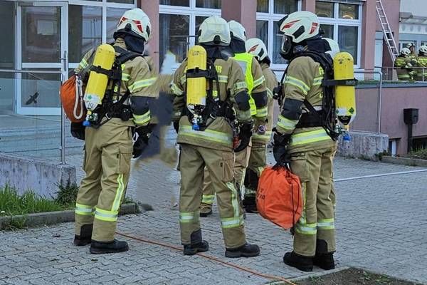 A group of firefighters in full gear, including helmets and oxygen tanks, are standing outside a building.