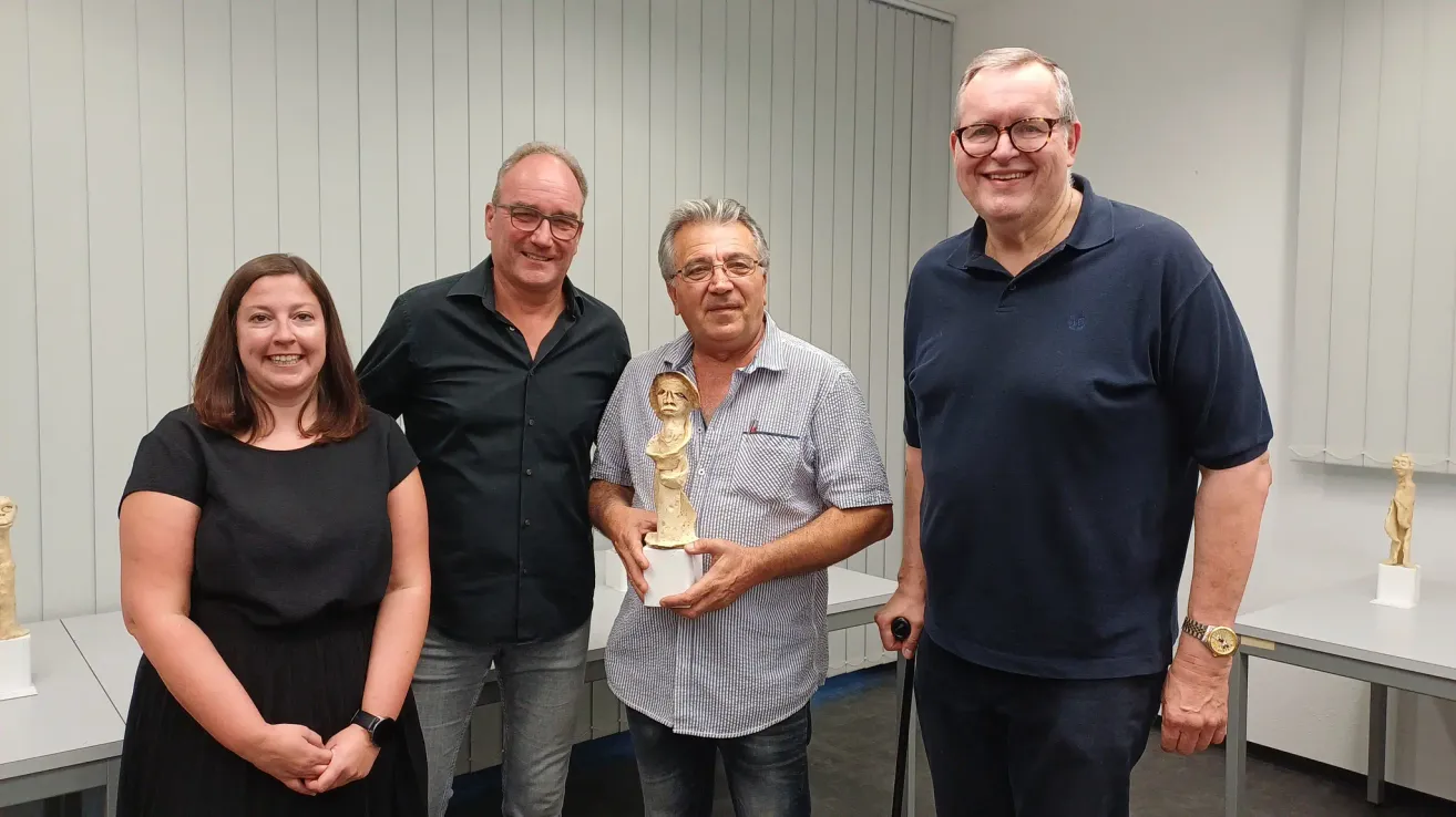 Four people are standing in a room, with one man holding a trophy. The man in the middle is wearing glasses and holding a golden trophy. The man on the right is holding a cane. The man on the left is wearing a watch.