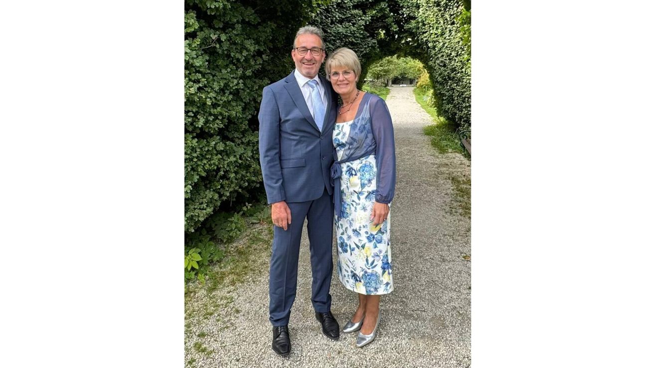 Ein Mann und eine Frau stehen auf einem Kiesweg und lächeln für ein Foto. Der Mann trägt einen blauen Anzug und die Frau ein Blumenkleid.