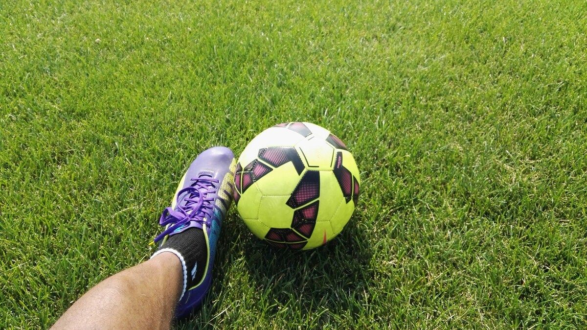 Ein Fußball mit einem gelben und schwarzen Muster liegt auf einem grünen Rasen neben einem Fuß, der einen lila Schuh trägt.