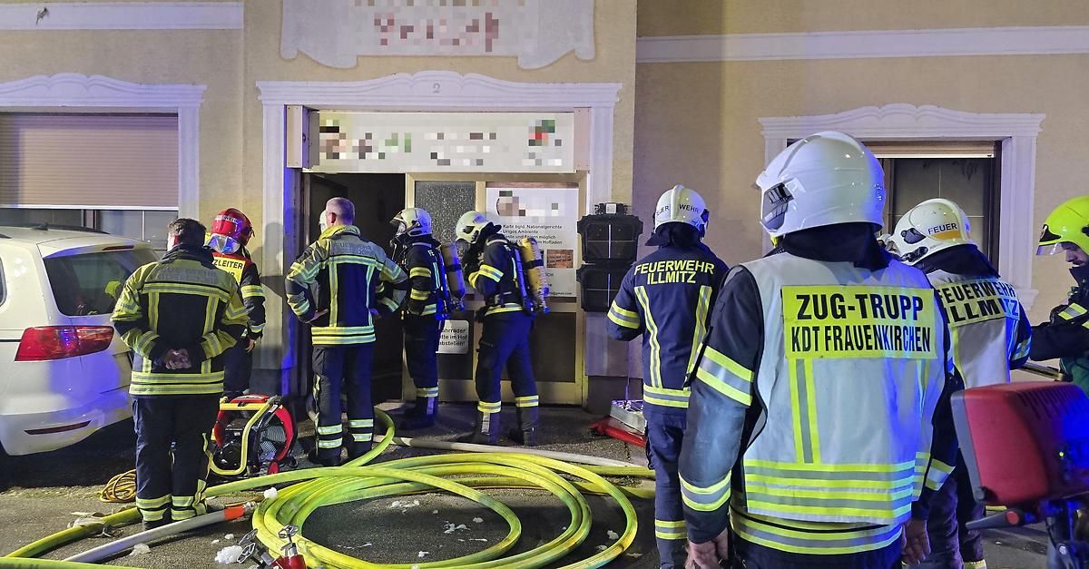 Eine Gruppe Feuerwehrleute in voller Ausrüstung steht vor einem Gebäude, wobei einer einen Schlauch hält und ein anderer einen Helm mit der Aufschrift 'Feuerwehr Illmitz' trägt. Das Gebäude hat eine beschädigte Tür und ein Schild mit verschwommenem Text.
