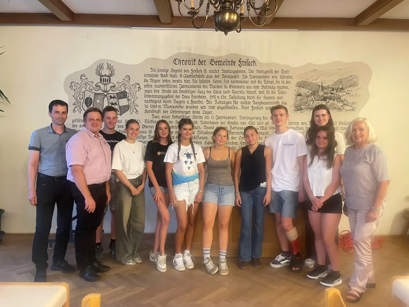 A group of people, including adults and teenagers, are posing for a photo in front of a mural depicting historical events and a coat of arms.