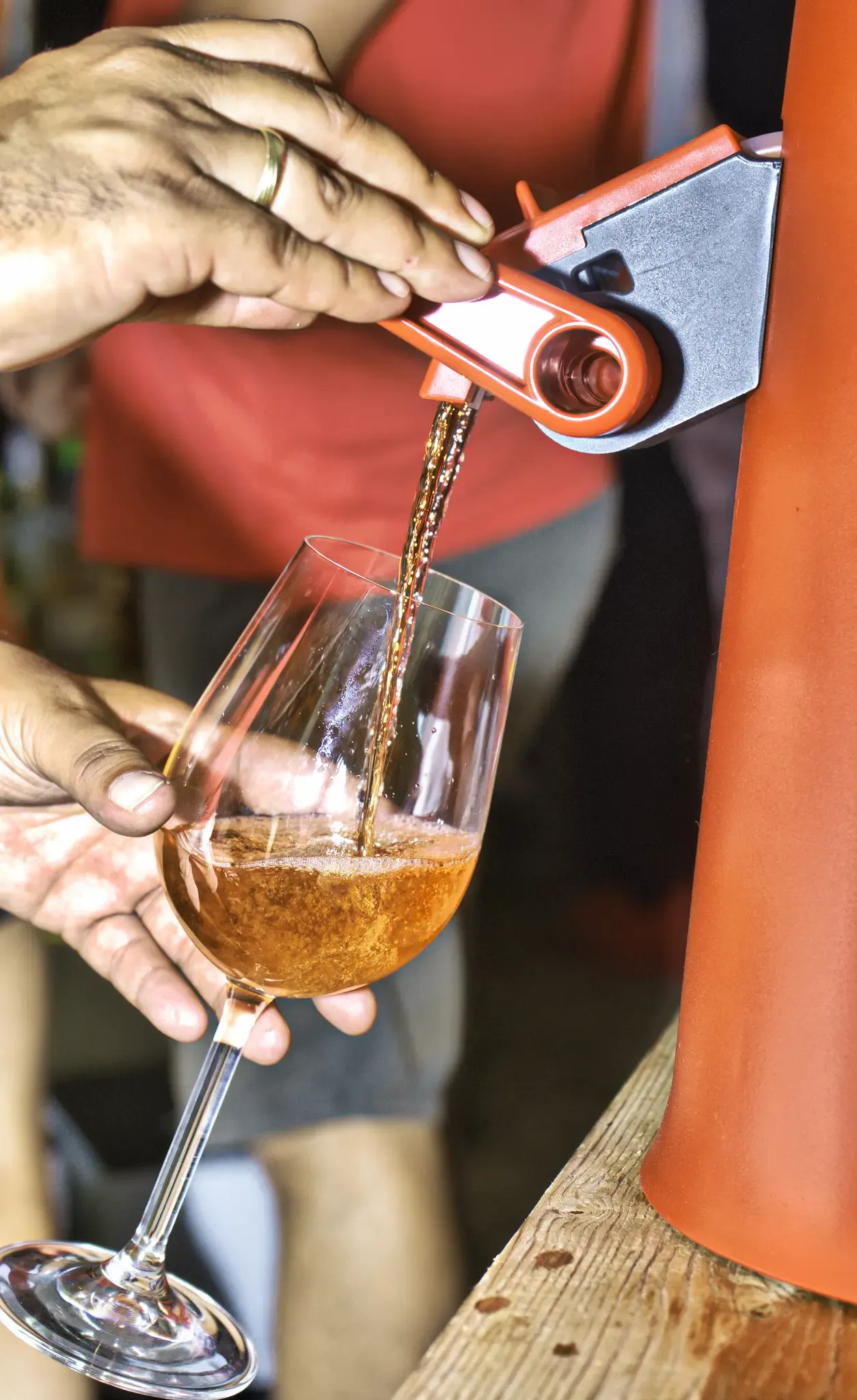 A person pours a golden liquid from a tap into a wine glass, with a blurry background.