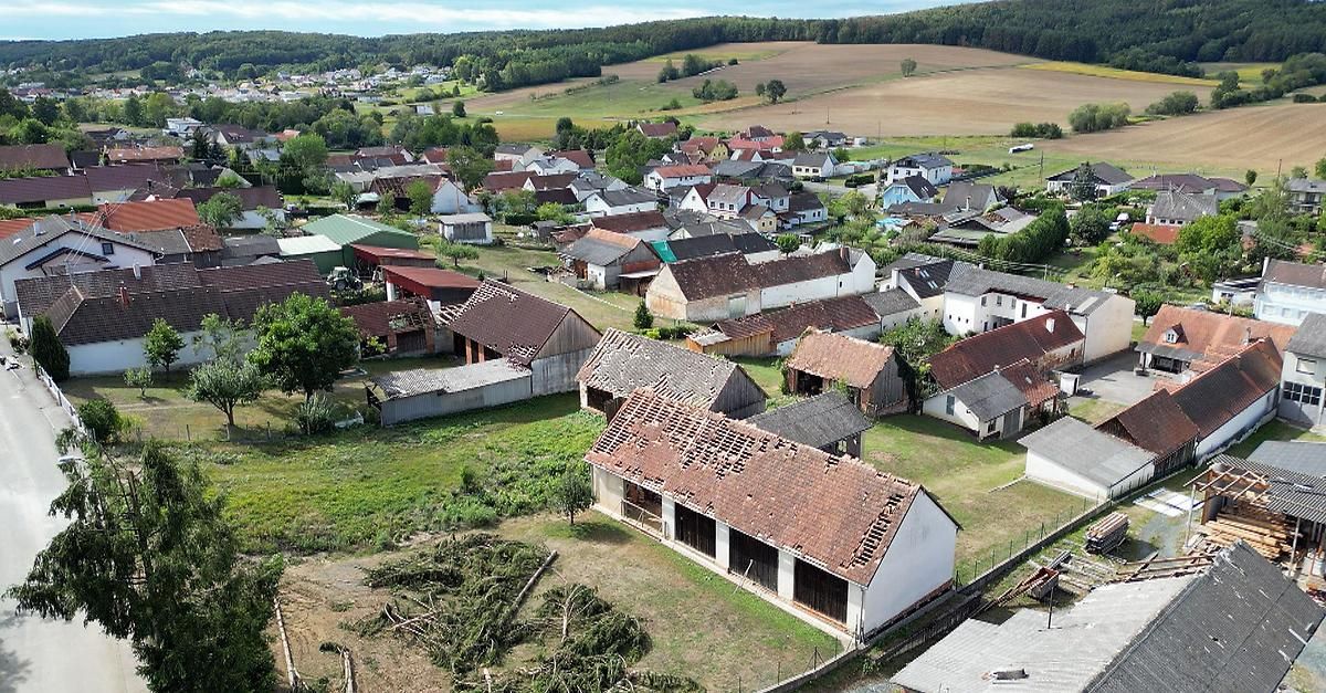 Luftaufnahme eines ländlichen Dorfes mit verschiedenen Häusern und Scheunen, einige in schlechtem Zustand, vor dem Hintergrund grüner Felder und weiter Hügel.