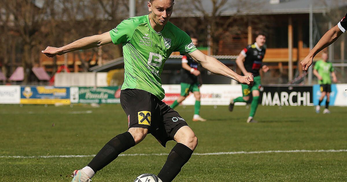 Ein Fußballspieler in einem grünen Trikot mit der Nummer 2 auf der Brust tritt einen Ball auf einem Feld. Dahinter laufen zwei weitere Spieler.