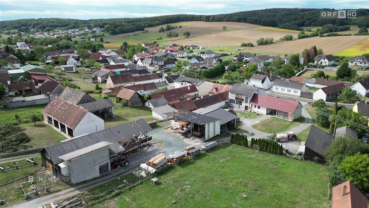 Luftaufnahme eines ländlichen Dorfes mit Häusern, Scheunen und einem Feld. Das Dorf ist von grünen Feldern und Bäumen umgeben.