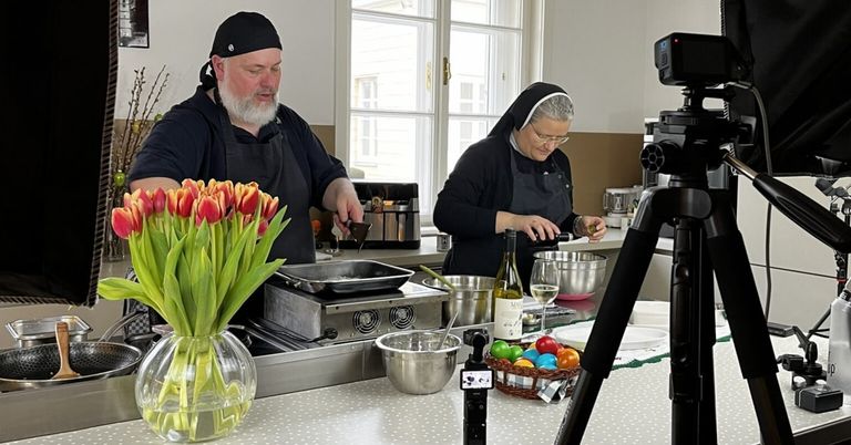 Ein Mann und eine Nonne kochen in einer Küche mit verschiedenen Küchenutensilien und Zutaten. Der Mann hält eine Schöpfkelle, und die Nonne gießt Wein in ein Glas. Es gibt Blumen, eine Flasche, einen Korb mit Eiern und ein Stativ mit einer Kamera in der Szene.
