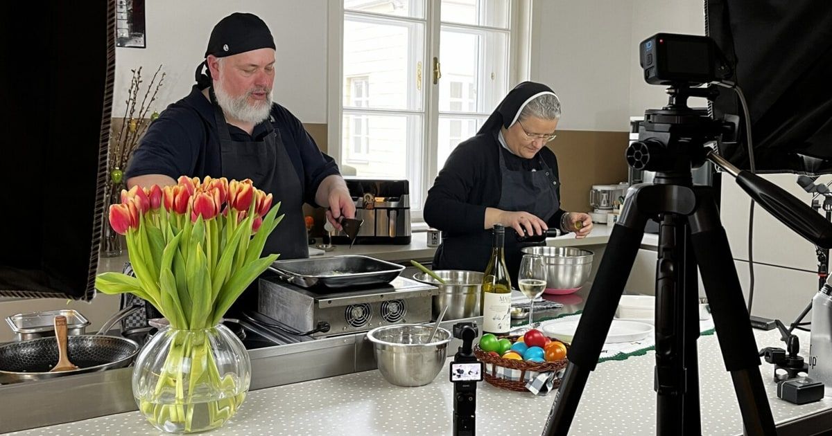 Ein Mann und eine Nonne kochen in einer Küche mit verschiedenen Küchenutensilien und Zutaten. Der Mann hält eine Schöpfkelle, und die Nonne gießt Wein in ein Glas. Es gibt Blumen, eine Flasche, einen Korb mit Eiern und ein Stativ mit einer Kamera in der Szene.