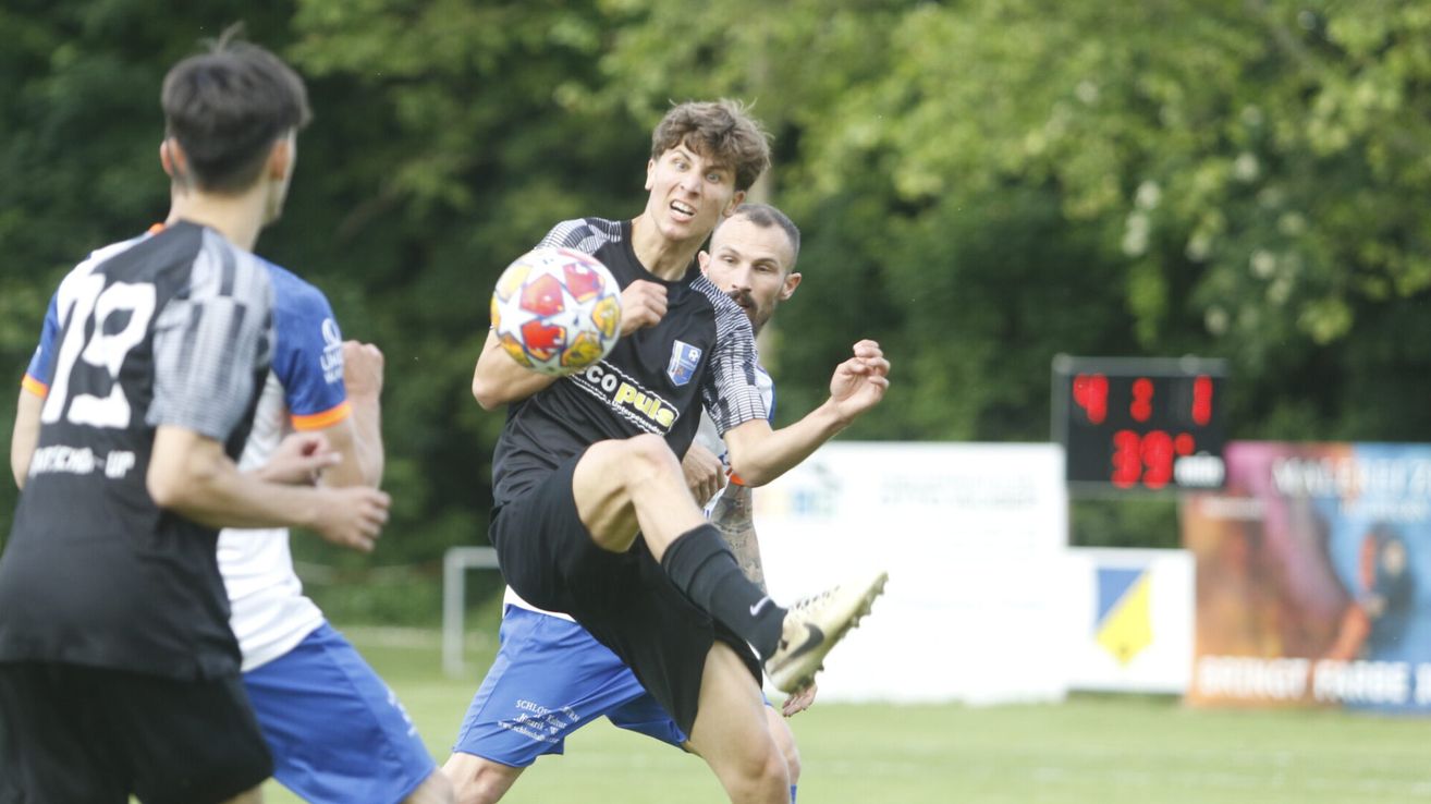 A soccer player in a black jersey with the word 'Copula' kicks the ball while another player in blue shorts tries to block him.