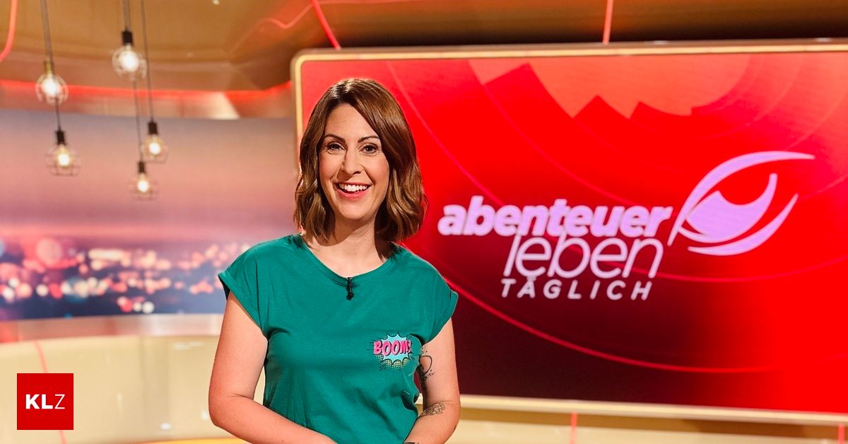 Eine Frau in einem grünen Shirt mit Logo steht lächelnd vor einem roten Schild mit weißem Text, das Abenteuer Leben TAGLICH steht.