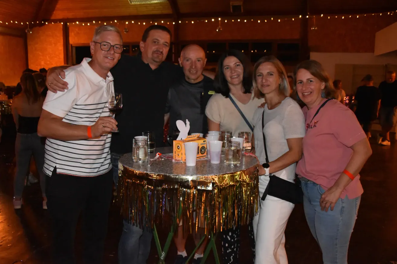 Eine Gruppe von sechs Erwachsenen, drei Männer und drei Frauen, posiert für ein Foto auf einer Party. Sie stehen um einen Tisch mit einem goldfarbenen Tischtuch mit Fransen, Gläsern und einer Schachtel mit Partyspielen. Der Raum ist mit Lichterketten dekoriert.