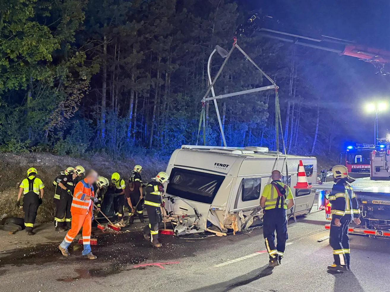Rettungskräfte räumen Trümmer von der Straße nach einem Unfall mit einem Wohnwagen. Der Wohnwagen wird von einem Kran angehoben.
