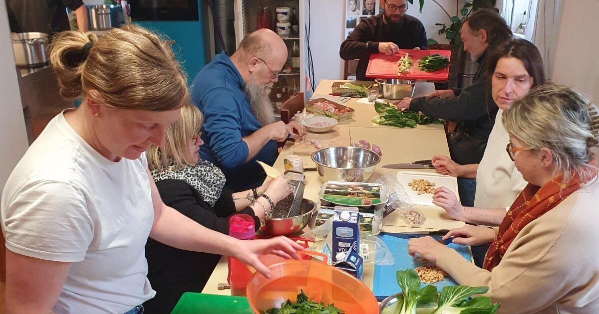 Eine Gruppe von Menschen ist um einen Tisch versammelt und bereitet Essen zu. Sie verwenden verschiedene Küchenutensilien und Zutaten.