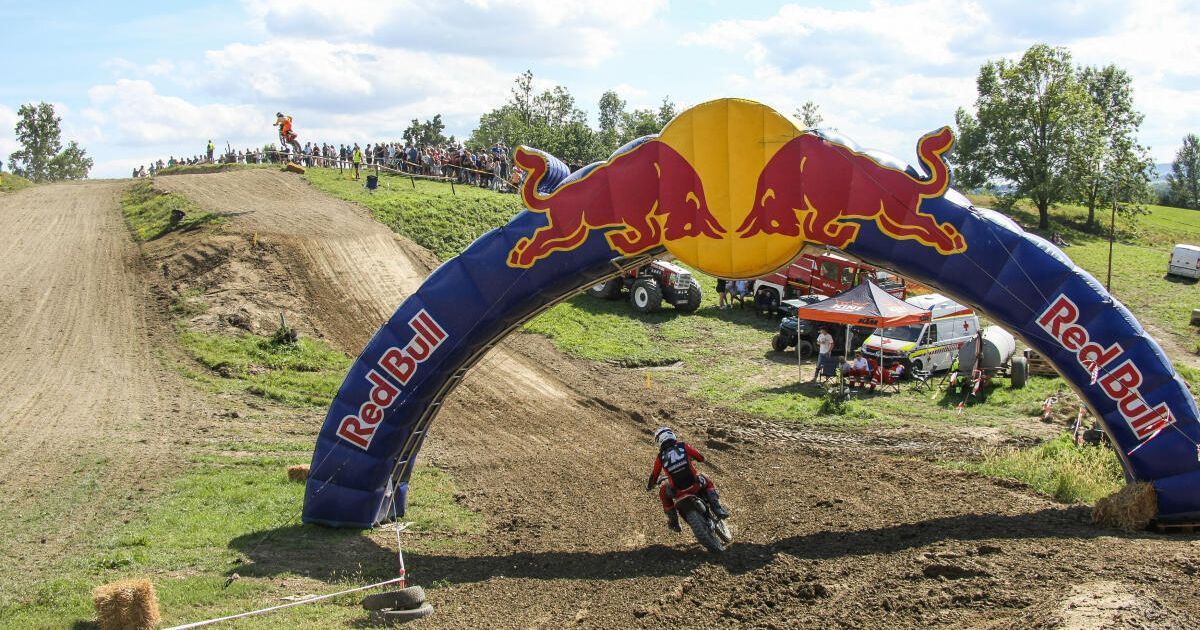 A motocross rider in red and white gear rides a motorcycle through a dirt track under a Red Bull archway. Spectators watch from the sidelines, and various vehicles and tents are set up nearby.