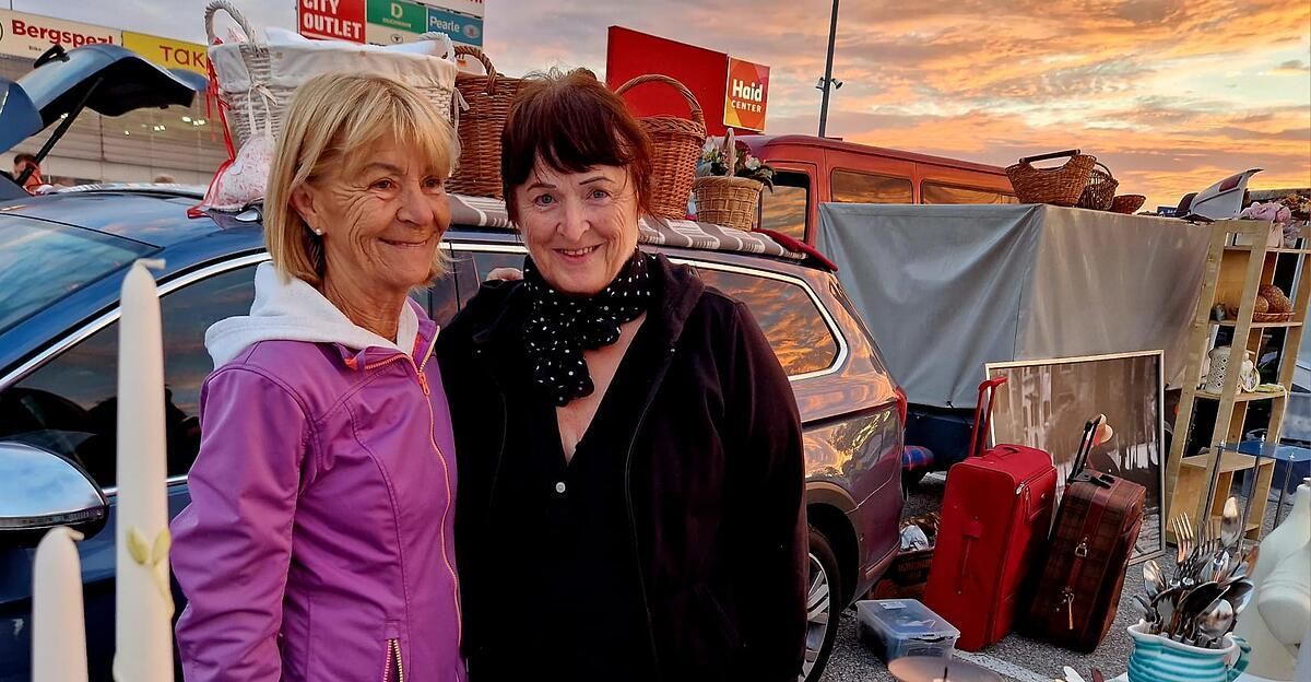 Zwei Frauen stehen lächelnd vor einem Auto mit Gepäck auf dem Dach. Dahinter sind ein Korb und ein Schild zu sehen, mit einem Sonnenuntergang im Hintergrund.