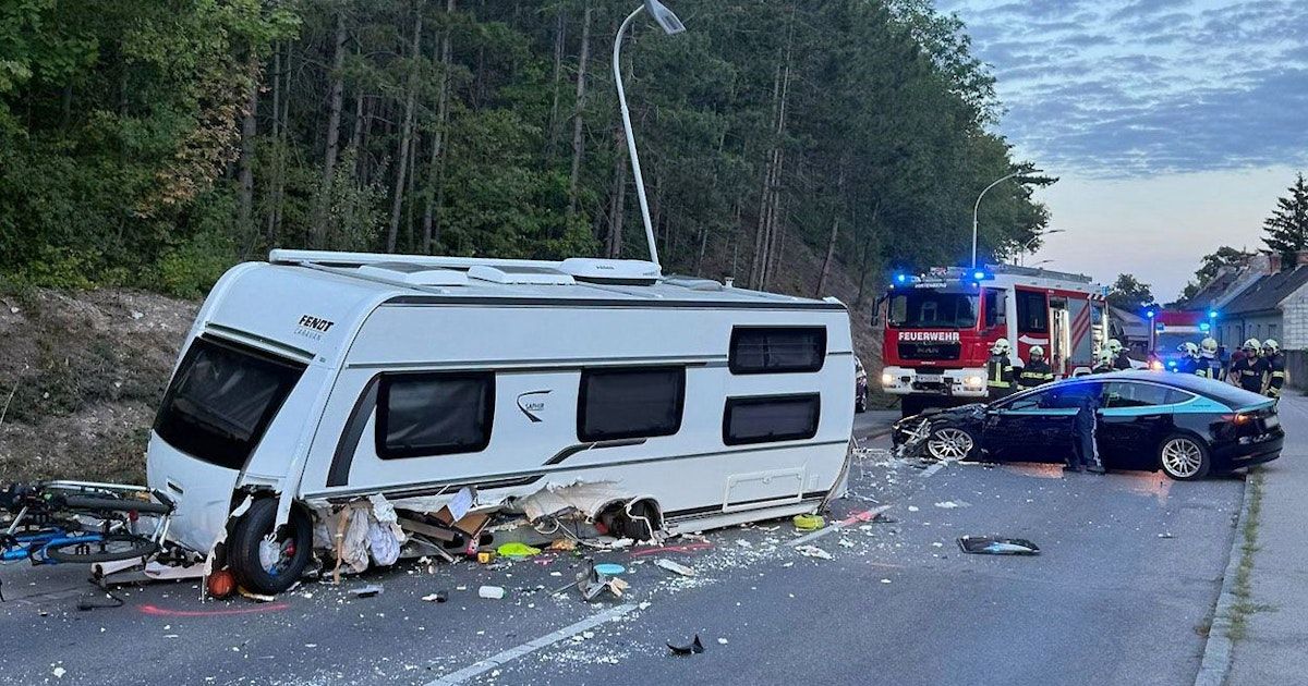 Ein weißer Wohnwagen ist auf der Straße umgekippt, mit herumliegenden Trümmern. Ein schwarzes Auto ist in der Nähe zerstört. Rettungsdienste sind anwesend, mit einem Feuerwehrwagen und einem Polizeiwagen.