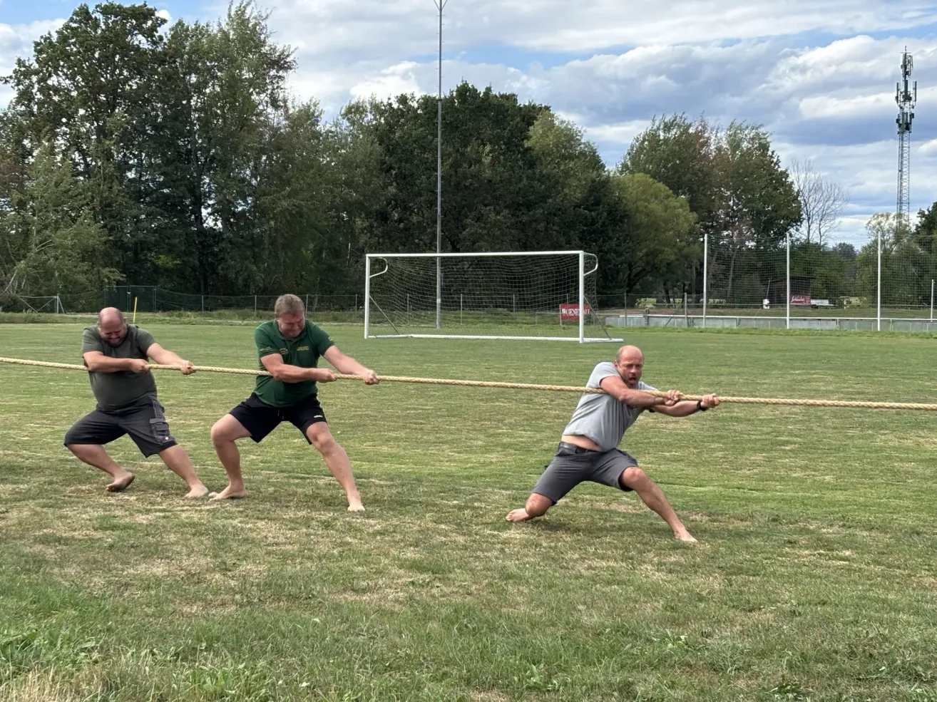 Drei Männer sind auf einem Rasenfeld in einem Tauziehen engagiert, mit einem Fußballtor im Hintergrund. Sie tragen alle lässige Sportbekleidung und scheinen sich auf das Spiel zu konzentrieren.