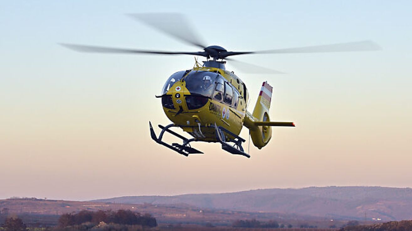 Ein gelber Hubschrauber mit roten Kreuzsymbolen an der Seite fliegt am Himmel. Der Hubschrauber hat eine Glaswindschutzscheibe und ein Fahrwerk. Der Himmel ist klar und blau.