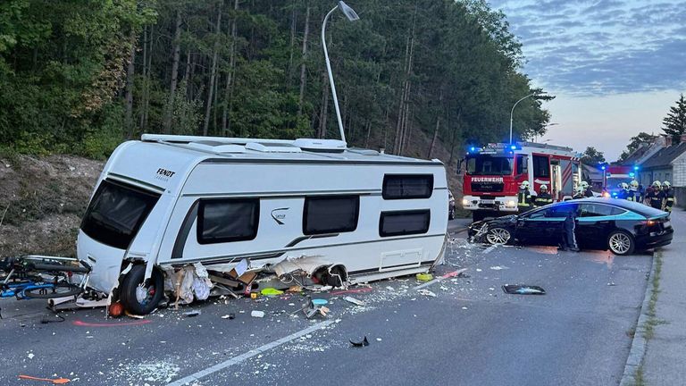 Ein weißer Wohnwagen ist auf der Straße umgekippt, mit herumliegenden Trümmern. Ein Feuerwehrwagen ist in der Nähe geparkt, und eine Person in einem gelben Weste steht daneben.