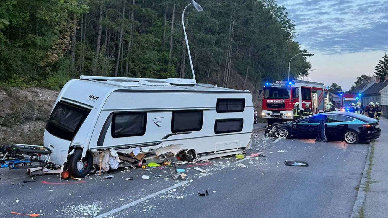 Ein weißer Wohnwagen ist auf der Straße umgekippt, mit herumliegenden Trümmern. Ein Feuerwehrwagen ist in der Nähe geparkt, und eine Person in einem gelben Weste steht daneben.