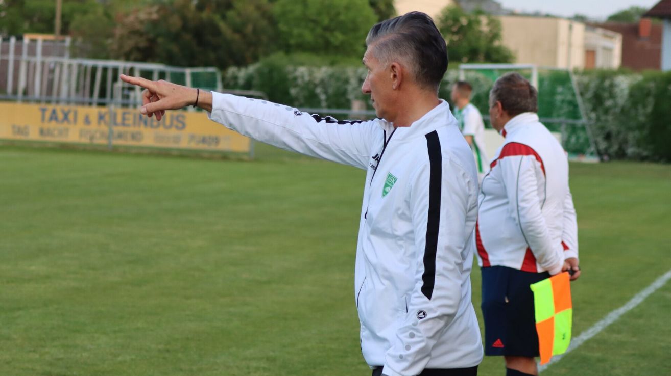 Ein Fußballtrainer in einer weißen Jacke mit grünen Akzenten zeigt auf das Feld, während zwei Spieler in weißen und roten Uniformen hinter ihm stehen.