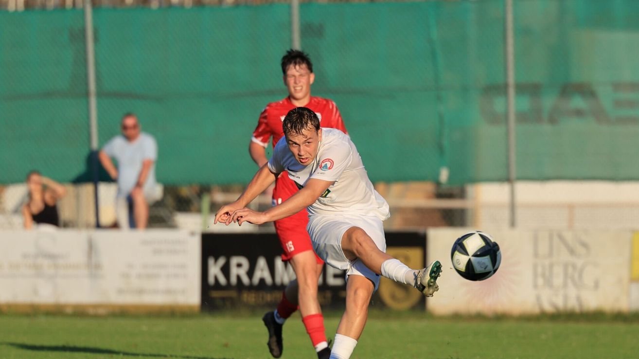 A soccer player in a white uniform is kicking the ball with his right foot while another player in red is chasing him.