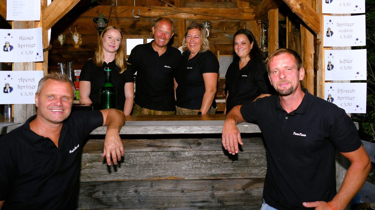 Eine Gruppe lächelnder Erwachsener in schwarzen Poloshirts steht hinter einer Holzbar, mit einem Mann rechts und einer Frau links, die eine Flasche hält.