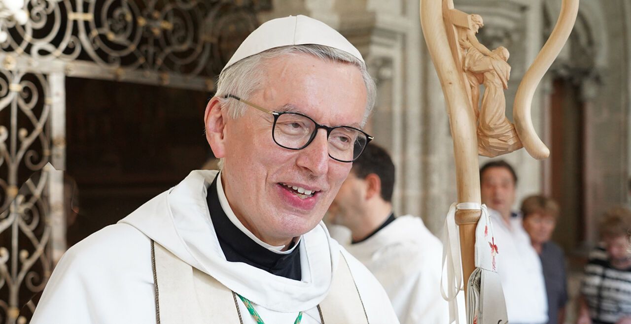 Ein lächelnder älterer Mann in einem weißen Hut und einer Brille steht in einer Kirche, mit einem hölzernen Kreuz und einer Statue hinter ihm.