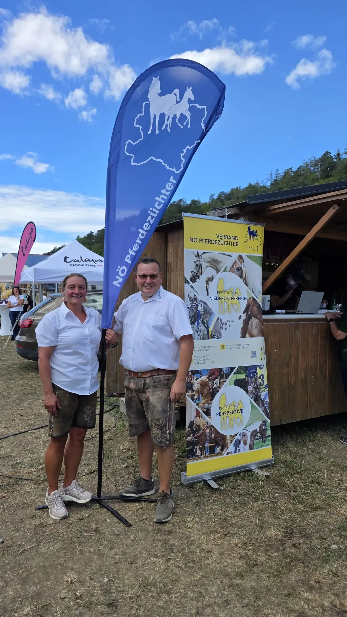 Zwei Personen stehen vor einem hölzernen Stand mit einem Banner. Der Mann trägt einen Gürtel und eine Sonnenbrille. Die Frau trägt ein weißes Shirt und Shorts. Hinter ihnen sind Zelte und ein Auto.