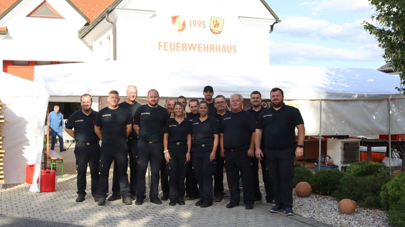 Eine Gruppe von Feuerwehrleuten in Uniform steht vor einem Gebäude mit der Aufschrift 'Feuerwehrhaus 1995' an der Wand. Sie posieren für ein Foto.