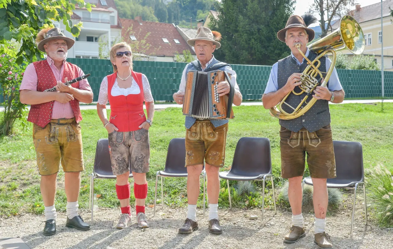 Vier Personen in traditioneller bayerischer Kleidung stehen auf einem Kiesweg, wobei zwei Männer Musikinstrumente spielen. Einer spielt Akkordeon, der andere Trompete. Eine Frau steht zwischen ihnen.
