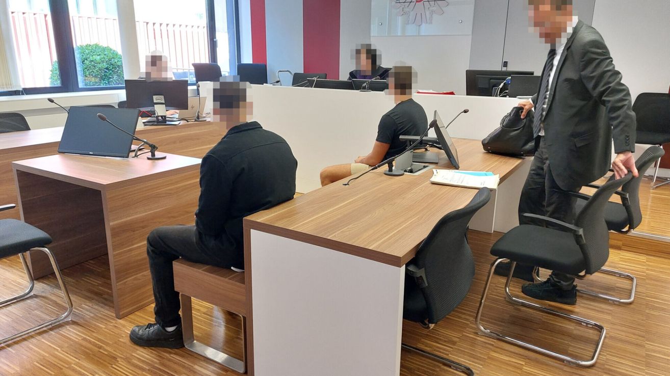 Zwei Personen sitzen in einer modernen Büroumgebung und sehen einen Schreibtisch mit Monitoren und Mikrofonen. Die Gesichter der Personen sind unkenntlich gemacht.