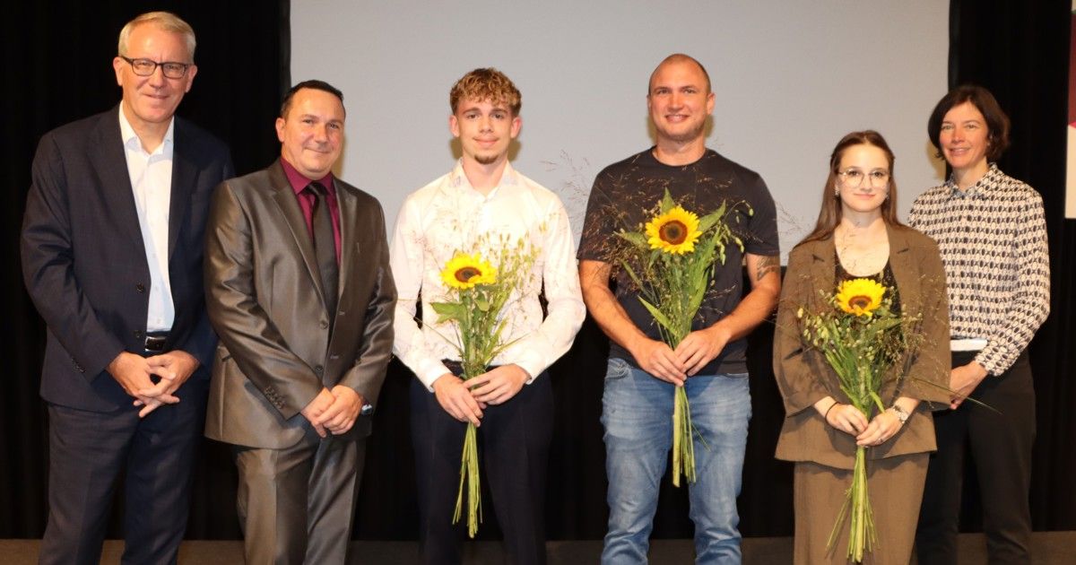 Four individuals stand together on a stage, smiling and holding sunflowers. The man on the left wears a suit, the second man a white shirt, the third man a black shirt, and the woman on the right wears a brown coat.