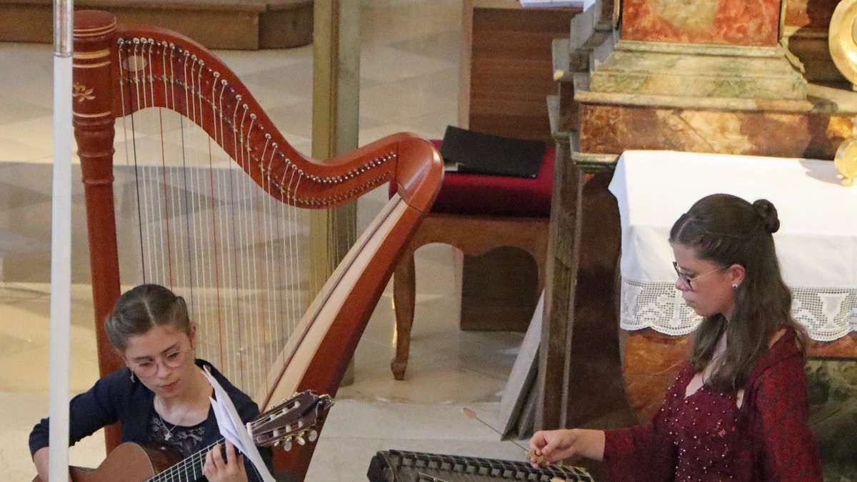 Zwei Frauen spielen Musikinstrumente in einer Kirche. Eine spielt die Harfe und die andere die Gitarre. Die Harfenistin konzentriert sich auf ihr Instrument, während die Gitarristin Notenblätter betrachtet. Dahinter steht ein Holzstuhl mit einem schwarzen Kissen und einem Buch.